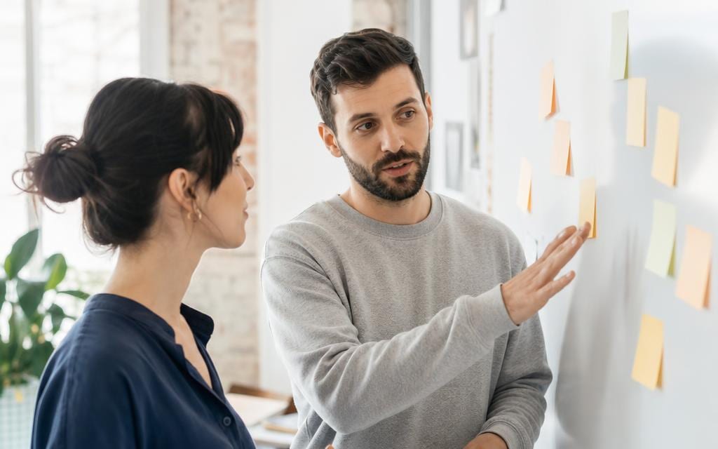 Two colleagues at an office whiteboard comparing ideas on blank sticky notes